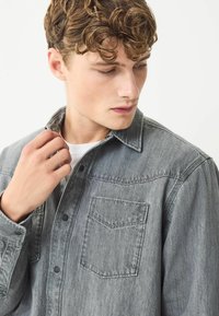 Grå denim skjorte med knaplukning, klassisk krave, frontlomme og subtil tekstur. Har sorte knapper og en afslappet pasform.