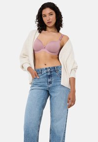Světle růžová podprsenka s vycpávkami a květinovými krajkovými detaily, nastavitelné ramínka a hladký povrch, v kombinaci se světle modrými džínami s vysokým pasem a krémovým kardiganem.