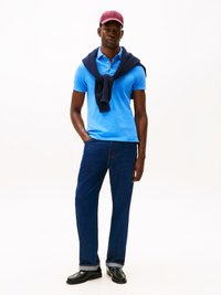 Camisola polo azul-claro com um logotipo, jeans azul-escuro, suéter azul-marinho amarrado sobre os ombros, e boné bordeaux. Sapatilhas pretas completam o visual.