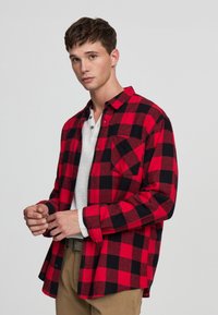 Giovane uomo che aggiusta le maniche indossando una camicia a scacchi rossa e nera sopra una t-shirt bianca e pantaloni khaki, in piedi contro uno sfondo chiaro e semplice.