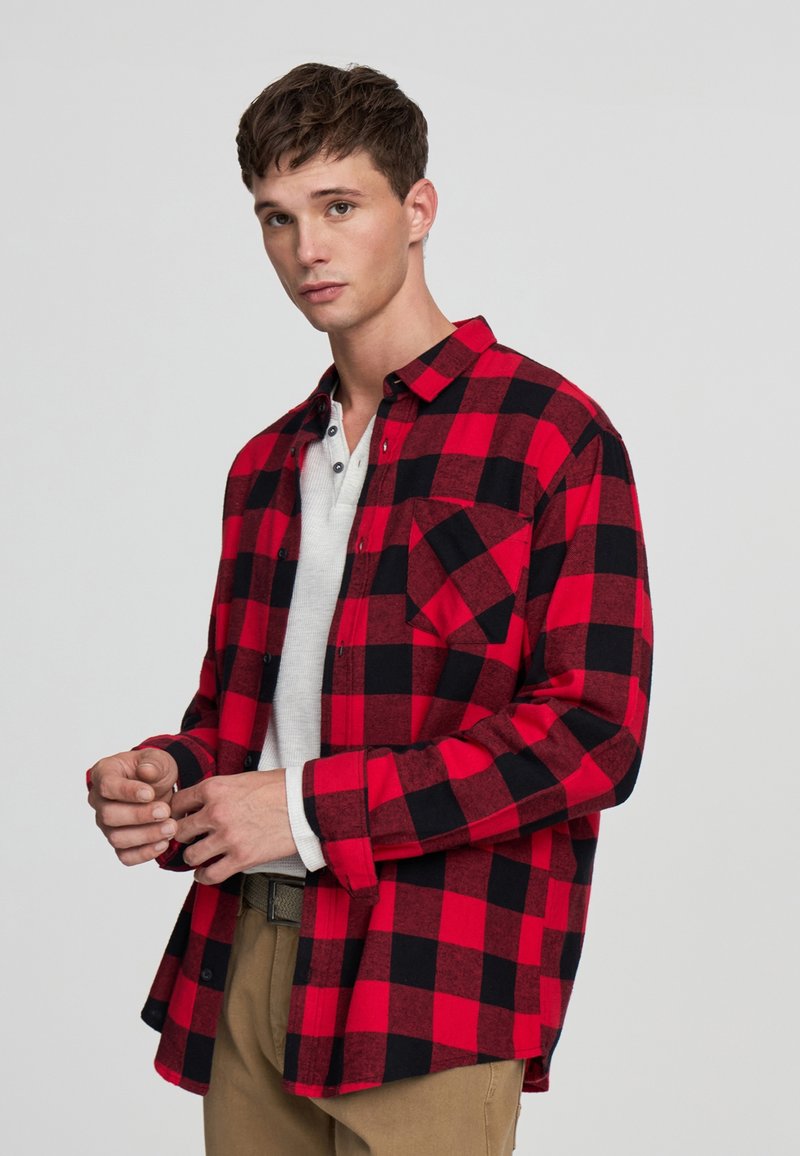 Giovane uomo che aggiusta le maniche indossando una camicia a scacchi rossa e nera sopra una t-shirt bianca e pantaloni khaki, in piedi contro uno sfondo chiaro e semplice.