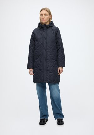 Femme portant un long manteau matelassé noir à capuche, un jean bleu à jambes larges et des chaussures noires épaisses, debout devant un fond blanc uni.
