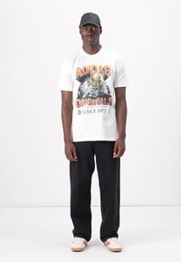 Hvid bomulds t-shirt med grafisk print og teksten "ADIDAS ORIGINALS", parret med sorte bukser og beige sneakers med røde detaljer.