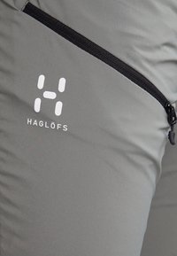 Šedé kalhoty z technické tkaniny s kapsou na zip a bílým logem ("H") s nápisem značky "HAGLÖFS" pod ním.