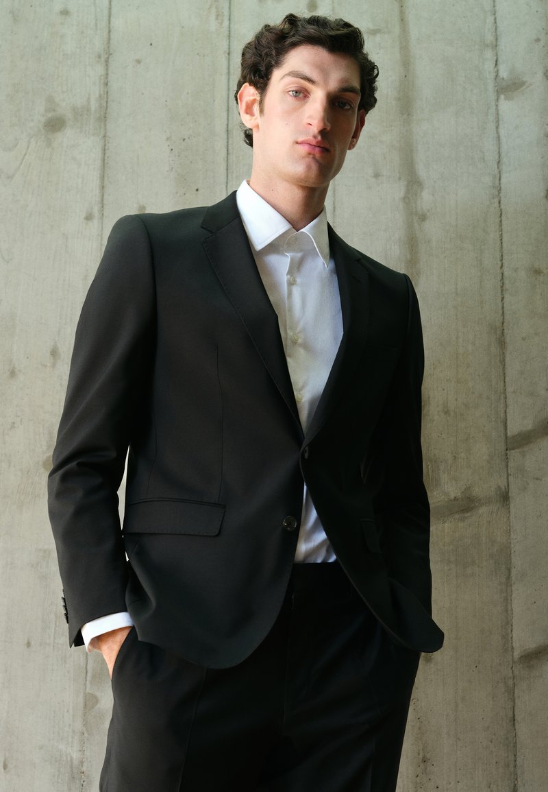 Traje negro con un diseño ajustado, que presenta un cierre de un botón, solapas con muesca y bolsillos laterales. Camisa blanca debajo, de pie contra una pared de concreto.