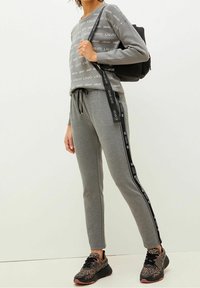 Szary sweatshirt z powtarzającym się nadrukiem logo, zestawiony z dopasowanymi dresami z czarnymi paskami po bokach. W zestawie z czarnymi i zwierzęcym wzorem sneakersami.