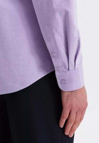 Camisa de color púrpura claro con puño de dos botones y una textura sutil, llevada sobre un fondo negro. Detalle de costura clara visible.