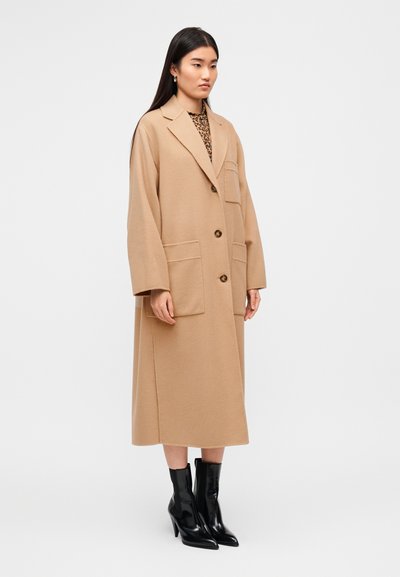 Μπεζ μακρύ παλτό με γιακά με χαραγή και δύο μπροστινές τσέπες, με κλείσιμο με ένα κουμπί και φαρδιά μανίκια, συνδυασμένο με μαύρα μποτάκια μέχρι τον αστράγαλο.