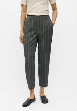 Person iført korte mørkegrå bukser med striber, beige kortærmet top og sorte slip-on sko, står med den ene hånd i lommen.