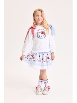 Νεαρή κοπέλα που φοράει φούτερ Hello Kitty με κόκκινες και μπλε φιόγκους, φούστα Hello Kitty ταιριαστή, κάλτσες και λευκά αθλητικά παπούτσια με κόκκινες κορδόνια.