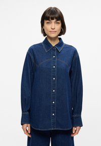 Τζιν πουκάμισο σε σκούρο μπλε με κουμπιά, μπροστινές τσέπες και αντίθεση ραφών. Διαθέτει κλασικό γιακά και μακριά μανίκια.