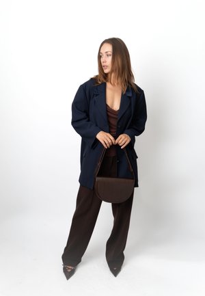 Sacou supradimensionat bleumarin, top maro, pantaloni maro și geantă maro texturată. Tocuri negre ascuțite completează ținuta. Fundal simplu.