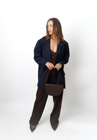 Blazer navy oversize, top marrone, pantaloni marroni e borsa testurizzata marrone. Décolleté nere a punta completano l'outfit. Sfondo semplice.