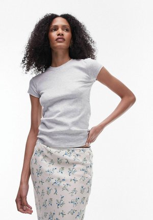 Γυναίκα με σγουρά μαλλιά που φοράει μια ανοιχτό γκρι εφαρμοστή μπλούζα και μια λευκή φούστα με μικρό μπλε ανθισμένο σχέδιο, στέκεται με το ένα χέρι στη μέση της.