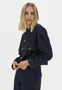 Tmavě modrá džínová bunda s cropped designem, která disponuje knoflíkovým zapínáním, předními kapsami a kontrastním oranžovým prošíváním. Měkká textura, uvolněný střih.