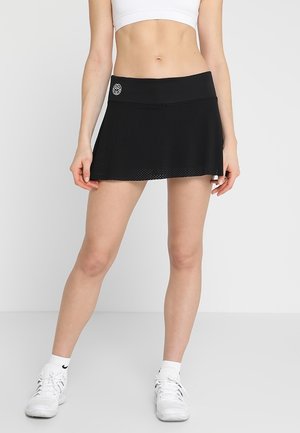 Vrouw draagt zwarte sportrok en witte sport-bh, staand met ontspannen handen langs het lichaam tegen een egale achtergrond.