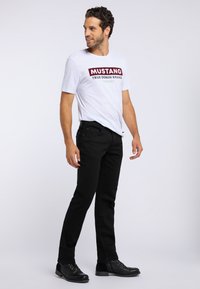 Biała koszulka z czerwonym nadrukiem "MUSTANG TRUE DENIM BRAND", w połączeniu z czarnymi dżinsami i czarnymi butami na sznurówki. Model stoi w profilu.