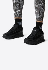 Svarte treningssko med en teksturert mesh-overside, med snøring og en solid gummisåle. Brukt med svarte ankelsokker og mønstrede leggins.