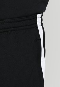 Schwarze Sporthose mit weißen Seitestreifen, aus einem weichen Stoff gefertigt, mit elastischem Bund und einer kleinen Tasche an der Seite.