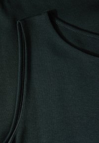 Tummanvihreä hihaton paita, joka on valmistettu pehmeästä materiaalista. Siinä on pyöreä pääntie ja pieni tasku edessä. Tasainen pinta.