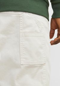 Pantaloni cargo di un leggero beige con una tasca posteriore e dettagli di cucitura visibili. Il tessuto appare morbido con una leggera consistenza. Indossati con un maglione verde.