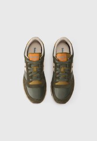 Zelene tenisice izrađene od kombinacije antilopa i mrežastih materijala. Imaju narančasti logo "Saucony JAZZ" te crne, smeđe i bež akcentne boje.