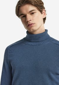 Blå stickad polotröja med ribbad krage och slät textur. Har en figurnära design och subtila detaljer på axlarna.