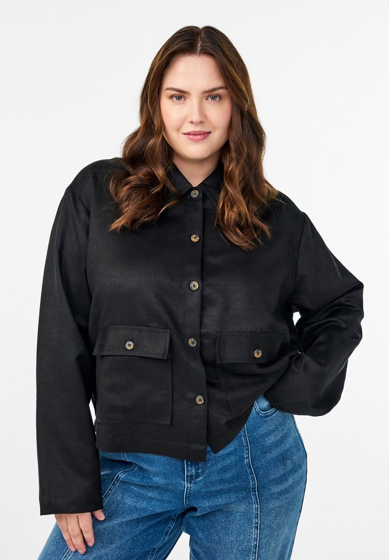 Donna con capelli castani ondulati indossa una giacca nera abbottonata con due tasche frontali e jeans blu a vita alta, posando con una mano sul fianco.