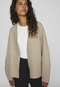 Cardigan beige avec une texture douce, à ouverture devant et épaules tombantes, porté sur une chemise blanche. Un pantalon noir complète l'ensemble.