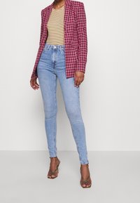 Person wearing a red plaid blazer, yellow striped top, light blue skinny jeans, and olive green high-heeled sandals standing against a white wall.