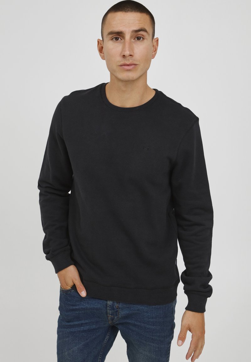 Felpa nera in cotone con colletto a giro, polsini e orlo a costine, caratterizzata da un piccolo dettaglio del logo sul davanti. Indossata con jeans blu.