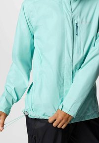 Giacca impermeabile di un leggero teal con tasca sul petto con zip, orlo regolabile con cordino e polsini elastici per una vestibilità sicura.