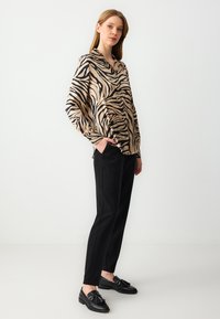 Beige blouse met zwarte zebra-strepen, lange mouwen, V-hals, gecombineerd met op maat gemaakte zwarte broek en zwarte instappers met kwastjes.