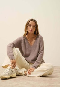Sweter w kolorze mauve z dekoltem w szpic, wykonany z miękkiej, strukturalnej tkaniny, w połączeniu z kremowymi spodniami o szerokiej nogawce i beżowymi sneakerami na neutralnym tle.