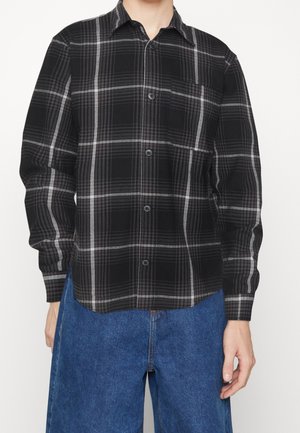 Homme portant une chemise à carreaux noire et grise à manches longues boutonée et un jean en denim bleu, debout devant un fond uni.