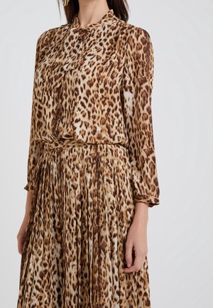 Kvinde iført en langærmet, brun og beige leopardprint bluse og matchende plisseret nederdel mod en ensartet lys baggrund.