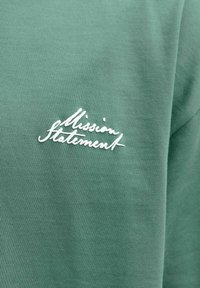 Roheline kangas valge kursiivse kirjaga "Missiooni avaldus" riideeseme rinnaosas tikitud.