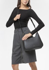 Borsa a mano in pelle nera con un design curvo, dotata di chiusura a zip e di una lunga tracolla regolabile, abbinata a una maglia nera aderente e a una gonna a righe grigie.