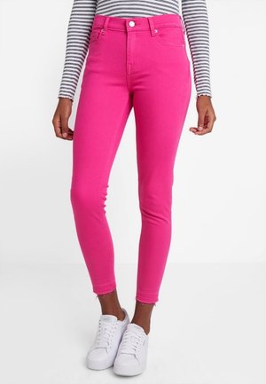 Osoba w jaskrawo różowych obcisłych dżinsach z surowym wykończeniem, białych trampkach i długim czarno-białym paskowanym topie, stojąca na białym tle.