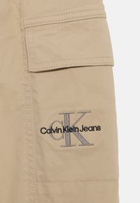 Beige nailonkangas, jossa on teksturoitu pinta, sisältää läppätaskun ja tummanharmaalla ja mustalla brodeeratun "Calvin Klein Jeans" -logon.