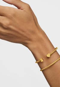Brazaletes en tono dorado en una muñeca, uno con un acento de corazón y el otro con un detalle de corazón más pequeño. Acabado metálico suave y diseño flexible.