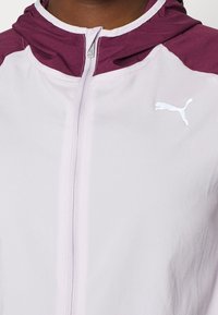 Veste zippée violet clair avec une capuche bordeaux, arborant un logo Puma blanc sur le côté gauche de la poitrine et une texture de tissu lisse.