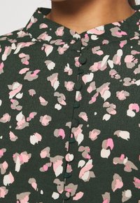 Grüne Bluse mit einem floralen Muster in Pink, Creme und Grau, mit Stehkragen und stoffbezogenen Knöpfen auf der Vorderseite.