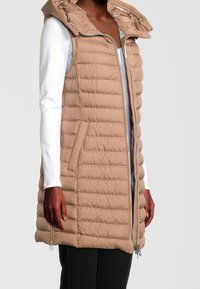 Chaleco acolchado beige con capucha, que cuenta con un cierre frontal, bolsillos laterales y detalles de costura. Llevado sobre una blusa blanca de manga larga.