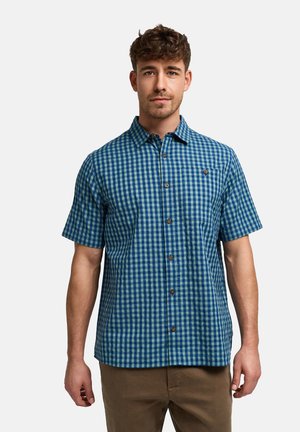 Mężczyzna ubrany w niebiesko-zieloną kraciastą koszulę z krótkim rękawem i brązowe spodnie, stojący na jednolitym tle.