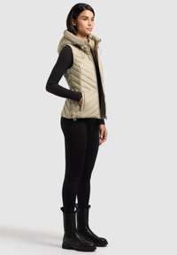 Femeie purtând vestă bej matlasată, fără mâneci, cu glugă, peste un top negru cu mâneci lungi, colanți negri și cizme negre până la jumătatea gambelor, stând în profil.
