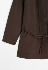 Bruine lange mouwen jas met voorvak en een dunne ceintuur gestrikt in de taille, weergegeven tegen een witte achtergrond.