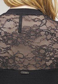 Top in pizzo nero con motivi floreali, schiena trasparente, bordo solido alle spalle e un insignia metallica "Guess" nella parte inferiore.