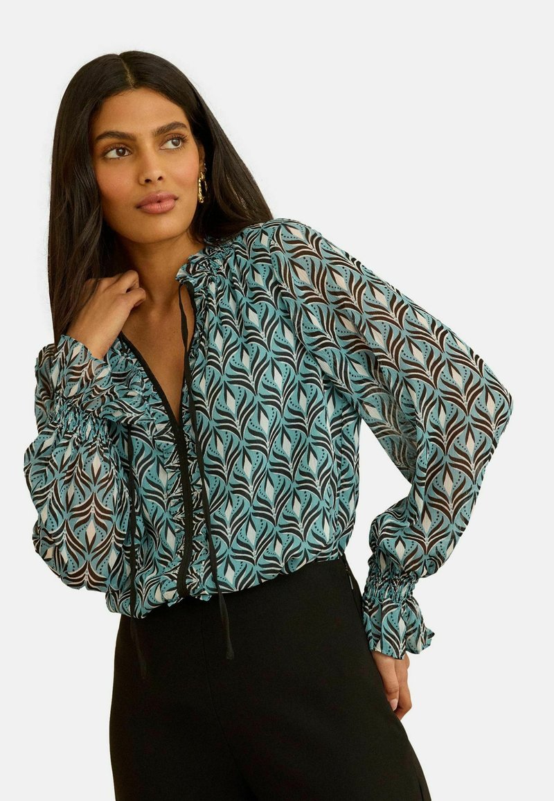 Žena s dugom tamnom kosom koja nosi plavuza u zeleno-crnoj uzorku s nabranim manžetama i crne hlače, zamišljeno gleda u stranu.