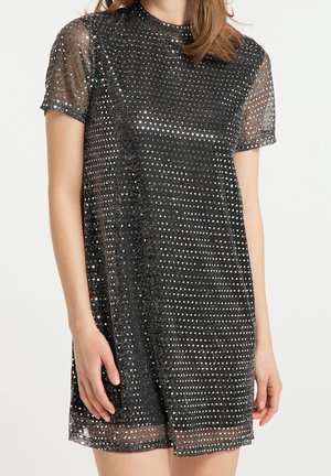 Femme portant une mini-robe noire à manches courtes avec un voile transparent recouvert de petits points argentés brillants.